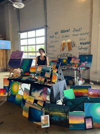 a woman standing in front of a table full of paintings